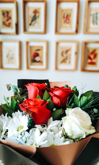 A floral arrangement featuring a bouquet of fresh red roses with velvety petals and vibrant green leaves, complemented by white flowers with delicate petals arranged in a blush pink wrapping paper. Th