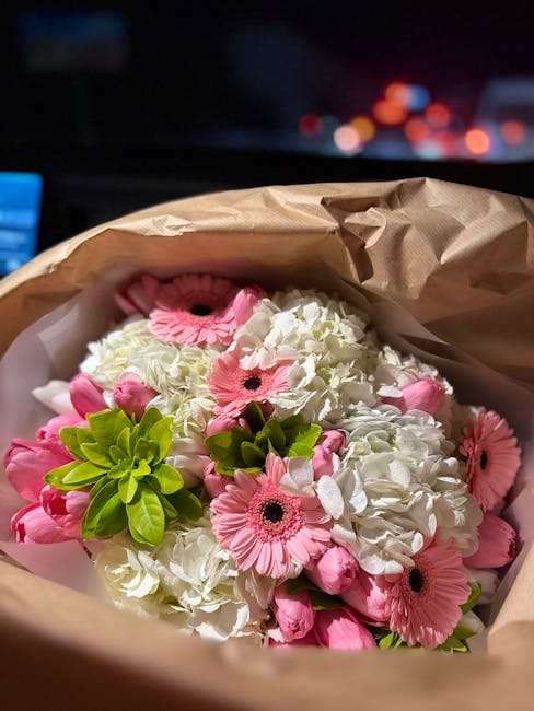 A floral bouquet arranged with white hydro chrysanthemums and pink gerbera daisies, accented by fresh green foliage. The flowers are wrapped in light brown kraft paper with a hint of white tissue pape