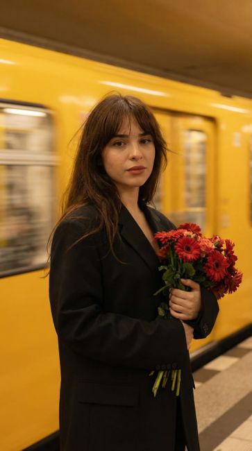 A young woman with long brown hair and bangs stands on a London underground platform, holding a bouquet of vibrant red and pink gerbera daisies arranged with green foliage. She is dressed in a black b